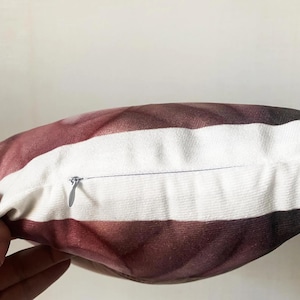 May include: Close-up of a decorative pillow with a striped pattern. The pillow features alternating bands of white and a gradient of burgundy to dark brown. A silver zipper is visible along the edge of the pillow.