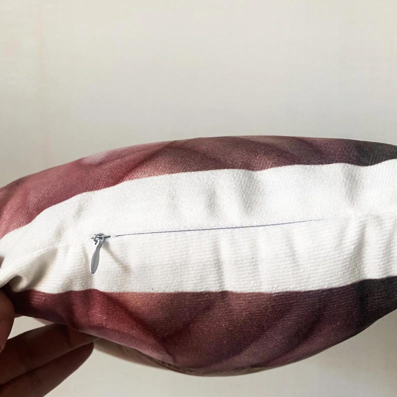 May include: Close-up of a decorative pillow with a striped pattern. The pillow features alternating bands of white and a gradient of burgundy to dark brown. A silver zipper is visible along the edge of the pillow.