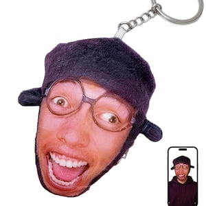 Op de afbeelding: Een novelty sleutelhanger met een afgedrukte afbeelding van een persoon met een bril en een hoed, met een wijd open mond. De sleutelhanger heeft een zilveren metalen ring en ketting. Een kleinere afbeelding van de persoon wordt weergegeven op een smartphone.