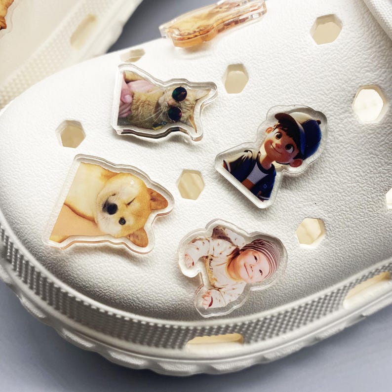 May include: White clogs with various photo charms attached. The charms include a cat, a dog, a baby, and a cartoon character.