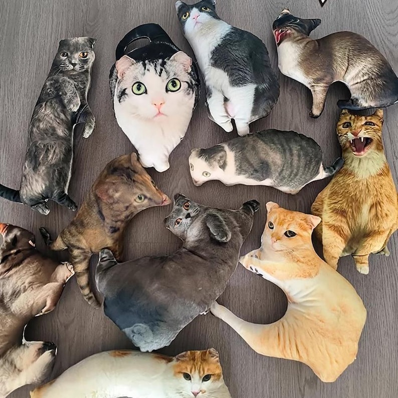 K&ouml;nnte beinhalten: Eine Sammlung von katzenf&ouml;rmigen Pl&uuml;schkissen in verschiedenen Farben und Mustern, darunter Tabby-, Calico- und Siam-Designs. Einige Kissen stehen, andere liegen. Die Kissen sind auf einer Holzoberfl&auml;che angeordnet.