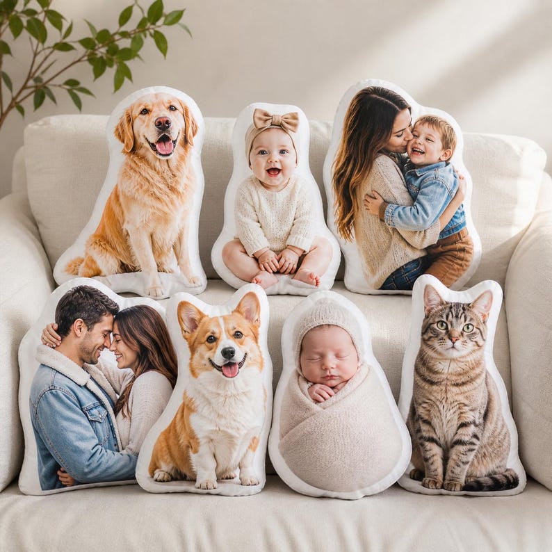 K&ouml;nnte beinhalten: Eine Sammlung von sieben individuell geformten Kissen mit aufgedruckten fotografischen Bildern eines Golden Retrievers, eines Babys mit Stirnband, einer Mutter mit Kind, eines Paares, eines Corgis, eines gewickelten Neugeborenen und einer getigerten Katze.