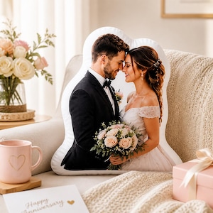 K&ouml;nnte beinhalten: Ein individuell geformtes wei&szlig;es Dekokissen mit einem aufgedruckten Foto eines Paares in Hochzeitskleidung. Das Bild zeigt eine Person im schwarzen Anzug und eine Person im wei&szlig;en schulterfreien Kleid mit einem Strau&szlig; aus rosa und wei&szlig;en Rosen.