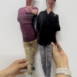 K&ouml;nnte beinhalten: Zwei Stoffpuppen, die jeweils einer Person &auml;hneln, werden vor einem wei&szlig;en Hintergrund gehalten. Eine Puppe tr&auml;gt ein lila T-Shirt mit Text, eine beige Hose und Turnschuhe. Die andere Puppe tr&auml;gt ein schwarzes Hemd und eine graue Hose. Beide Puppen haben die Nummer "27375" auf ihren Beinen aufgedruckt.