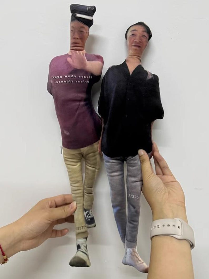 K&ouml;nnte beinhalten: Zwei Stoffpuppen, die jeweils einer Person &auml;hneln, werden vor einem wei&szlig;en Hintergrund gehalten. Eine Puppe tr&auml;gt ein lila T-Shirt mit Text, eine beige Hose und Turnschuhe. Die andere Puppe tr&auml;gt ein schwarzes Hemd und eine graue Hose. Beide Puppen haben die Nummer "27375" auf ihren Beinen aufgedruckt.