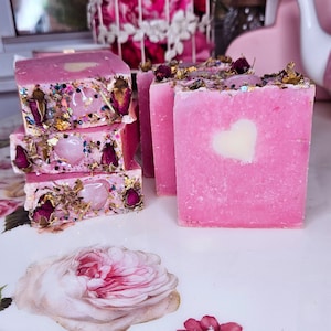 May include: Four pink soap bars with a heart design and embedded crystals and dried flowers. The soap bars are stacked on top of each other.