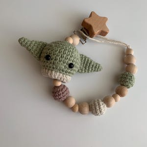 Op de afbeelding: Een gehaakt groen Baby Yoda-personage bevestigd aan een houten ster en een reeks houten en gehaakte kralen. De kralen zijn in tinten bruin, beige en roze.