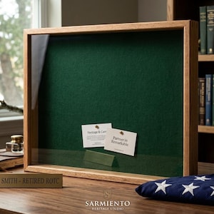 May include: A wooden display case with a green felt interior and a glass front. Two cards are pinned inside. A folded American flag and a nameplate that reads "COL. SMITH - RETIRED ROTC" are on the table. The case is from Sarmiento Heritage Studio.
