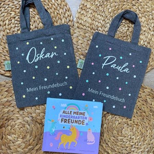 Könnte beinhalten: Zwei graue Stofftaschen mit weißen Punkten und den Namen "Oskar" und "Paula" darauf gedruckt. Die Taschen haben den Text "Mein Freundbuch" aufgedruckt. Ein Buch mit einer Regenbogen-, Einhorn- und Katzenillustration befindet sich in der Mitte des Bildes. Der Buchtitel lautet "Alle Meine Kindergarten Freunde".