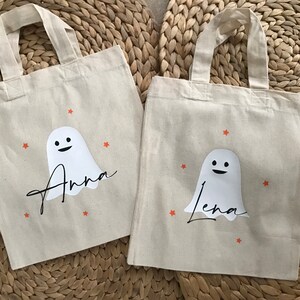 Könnte beinhalten: Zwei weiße Stofftaschen mit orangefarbenen Sternen und einem weißen Geist mit einem lächelnden Gesicht. Die Taschen sind mit den Namen "Anna" und "Lena" in schwarzer Kursivschrift personalisiert.