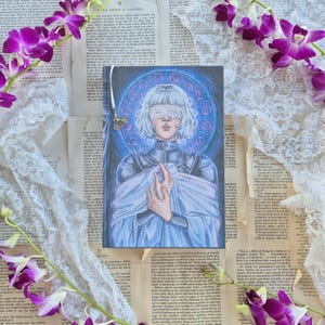 May include: A fantasy art print featuring a figure with a blindfold, set against a blue and white circular design. The artwork is on a book cover, surrounded by vintage book pages, lace, and purple orchids.