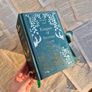 Pode incluir: Um livro de capa dura azul-esverdeado com letras douradas e ilustrações. O título é "Empire of Storms" e o subtítulo é "A Tower of Dawn". O livro faz parte de uma série de "leitura em tandem".