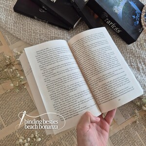 Puede incluir: Un libro abierto con p&aacute;ginas impresas, sostenido por una mano, con el texto visible. Varios libros negros con la palabra "Fearless" y "Powerful" est&aacute;n en el fondo. El texto "binding besties beach bonanza" est&aacute; en la esquina inferior izquierda.