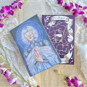 May include: Two books with illustrated covers. One features a figure with a blindfold in a blue and purple circular design. The other book is purple with gold text that reads "The Knight & the Moth Book 1".