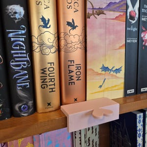 Könnte beinhalten: Ein rosa Buchstütze mit herzförmigem Design steht auf einem Holzregal neben einer Reihe von Büchern. Die Bücher tragen Titel wie "Nightbane", "Fourth Wing" und "Iron Flame". Die Buchumschläge sind in verschiedenen Farben gehalten.