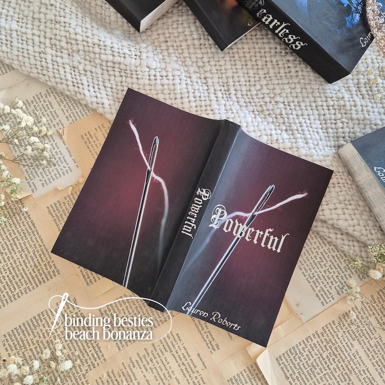 Puede incluir: Un libro abierto titulado "Powerful" con un dise&ntilde;o de aguja e hilo en la portada, sobre un fondo burdeos. El libro est&aacute; rodeado de otros libros y papel vintage. El texto "binding besties beach bonanza" tambi&eacute;n es visible.
