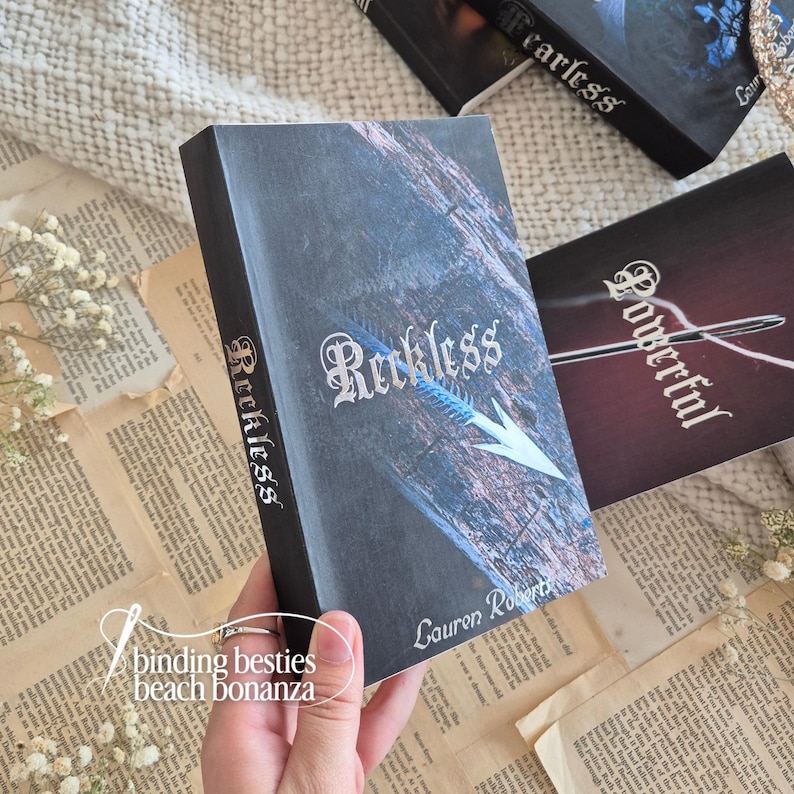 Puede incluir: Una pila de libros con cubiertas oscuras. El libro principal se titula "Reckless" con un dise&ntilde;o de flecha plateada. El nombre del autor, Lauren Roberts, es visible. Otros libros son visibles con los t&iacute;tulos "Fearless" y "Powerful". Las palabras "binding besties beach bonanza" tambi&eacute;n son visibles.