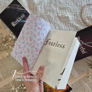 Puede incluir: Un libro abierto con la palabra "Fearless" en escritura negra se sostiene abierto, revelando guardas con estampado floral. Se ven otros libros con las palabras "Reckless" y "Powerful". La imagen incluye el texto "binding besties beach bonanza".