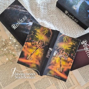 Puede incluir: Varios libros con cubiertas oscuras se exhiben, con t&iacute;tulos como "Fearful" y "Reckless". El libro abierto muestra un dise&ntilde;o de llave dorada sobre un fondo borroso y oto&ntilde;al. Tambi&eacute;n se ve el texto "binding besties beach bonanza".