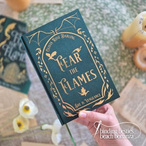 May include: A teal book with gold lettering and accents, featuring the title "Fear the Flames." The cover includes illustrations of dragons and foliage. The author's name, Olivia Rose Darling, is also displayed. A green ribbon bookmark is visible.