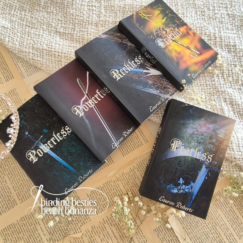 Puede incluir: Una colecci&oacute;n de libros de tapa dura con t&iacute;tulos como "Powerless", "Fearless" y "Reckless". Las portadas de los libros presentan ilustraciones de una espada, una llave y una aguja. El nombre del autor, Lauren Roberts, est&aacute; impreso en cada libro. Tambi&eacute;n se ve la frase "binding besties beach bonanza".