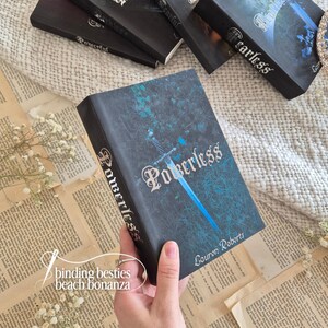 Puede incluir: Un libro de tapa dura titulado "Powerless" de Lauren Roberts, con un dise&ntilde;o de portada azul y negro con una ilustraci&oacute;n de espada. El libro se sostiene sobre p&aacute;ginas de libros antiguos, con otros libros visibles en el fondo.
