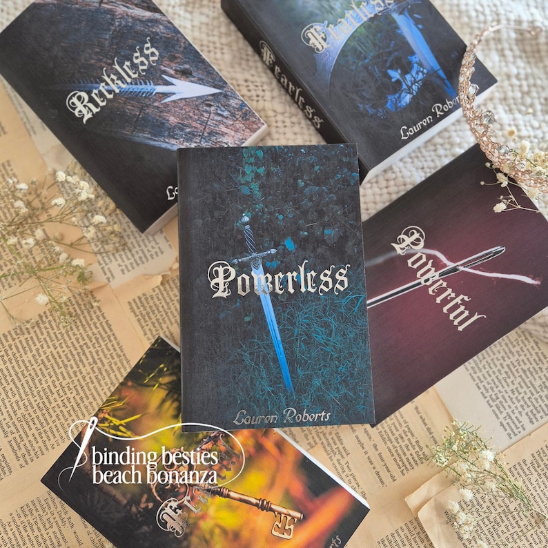 Puede incluir: Una colecci&oacute;n de novelas de fantas&iacute;a con t&iacute;tulos como "Reckless", "Fearless", "Powerless" y "Powerful". Las portadas presentan texto e im&aacute;genes estilizadas, incluyendo una espada y una flecha. Los libros est&aacute;n dispuestos sobre un fondo de papel envejecido y acentos florales.