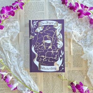 May include: A purple book titled "The Knight & the Poets" by Rachel Gillig, featuring a gold foil design of a woman with a blindfold and abstract patterns. The book is surrounded by vintage pages, lace, and purple orchids.