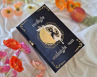 May include: A black book titled Twilight Midnight Sun with gold foil lettering and a central circular graphic featuring a split design of a sun and a pomegranate. The cover includes decorative gold corner accents and a small gold metal charm attached.