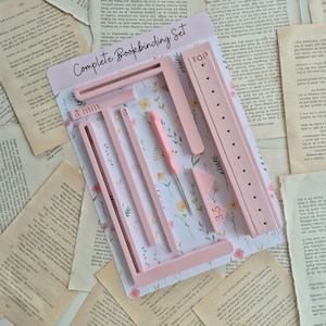 May include: A pink plastic bookbinding set with tools for sewing and stitching book pages. The set includes a ruler, a guide, a stitching awl, and a needle. The set is labeled "Complete Bookbinding Set" and includes measurements in millimeters.