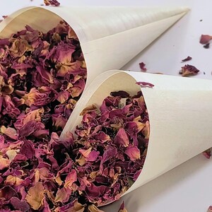 May include: Two off-white paper cones filled with dried rose petals in shades of deep magenta, burgundy, and muted pink. A loose pile of the same dried flower petals is scattered to the left of the cones, creating a textured floral display.