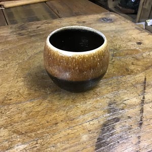 May include: A ceramic cup with a rounded shape, featuring a gradient of colours. The top rim is white, transitioning to a speckled brown, and ending with a dark brown base. The interior is black.