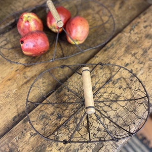 May include: Two oval wire baskets with wooden handles. One basket holds four red and yellow apples. The baskets are made of dark metal and rest on a rustic wooden surface, evoking a farmhouse style.