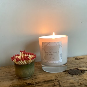 May include: A burning white soy candle, labelled "Sandalwood + Pepper", and a small green ceramic cup filled with red-tipped matches. The candle's flame glows, lighting the scene on a wooden surface.