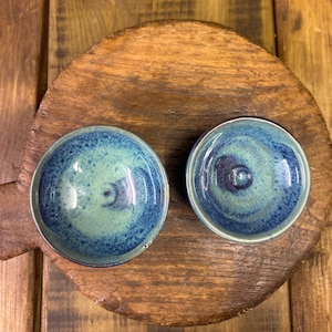 May include: Two small, round ceramic bowls with a glossy, speckled blue and green glaze. The bowls are set on a rustic, wooden chopping board, creating a contrast of textures and colours. The bowls have a small, circular indentation in the centre.
