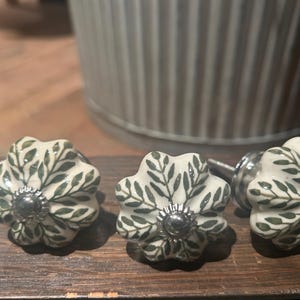 May include: Three floral cabinet knobs. Each knob has a white base with green leaf patterns and a silver centre. The knobs are displayed on a wooden surface.