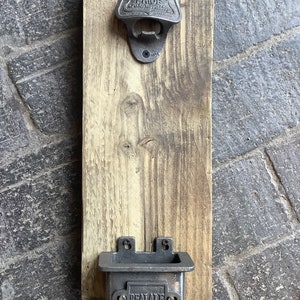 May include: A wall-mounted bottle opener and cap catcher on a wooden plank. The metal opener is marked "Fuller's London Pride". The cap catcher below has "Real Ale" text. The wood has a weathered look.