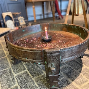 May include: A circular, antique wooden coffee table with a dark brown finish and decorative carvings. A small black bowl holds a lit pink candle, surrounded by rose petals. The table has a low profile and sturdy legs.