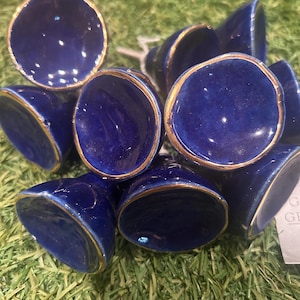 May include: A cluster of deep blue ceramic bells with gold edging. The bells are displayed on artificial green grass. The bells have a glossy finish and a rounded shape.
