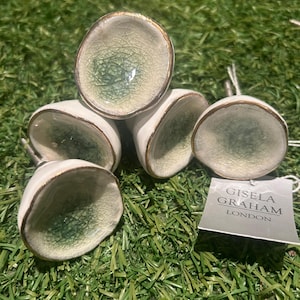 May include: A set of ceramic cabinet knobs. Each knob has a white exterior, a crackled green interior, and a gold-coloured rim. The knobs are displayed on green grass. A tag reads "GISELA GRAHAM LONDON".