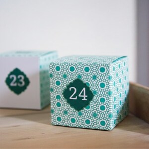 May include: Two cube-shaped boxes, one with the number 23 and the other with 24, are displayed on a wooden surface. The box with 24 has a teal and white geometric pattern. The numbers are in a dark teal diamond shape.
