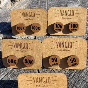 Puede incluir: Seis pares de pendientes redondos de madera con diferentes inscripciones. Las inscripciones incluyen "100K", "50K", "100 millas", "50 millas" y "ultra". Los pendientes se muestran en pequeñas tarjetas de madera con el nombre de la marca "VANGLO creations" impreso en ellas.