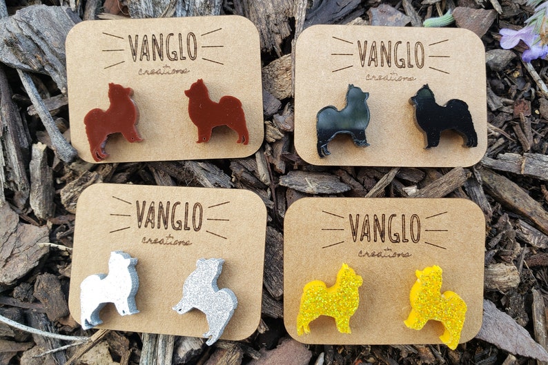 May include: Four pairs of dog silhouette earrings on brown cards. The earrings are made of acrylic and come in brown, black, silver, and yellow glitter.