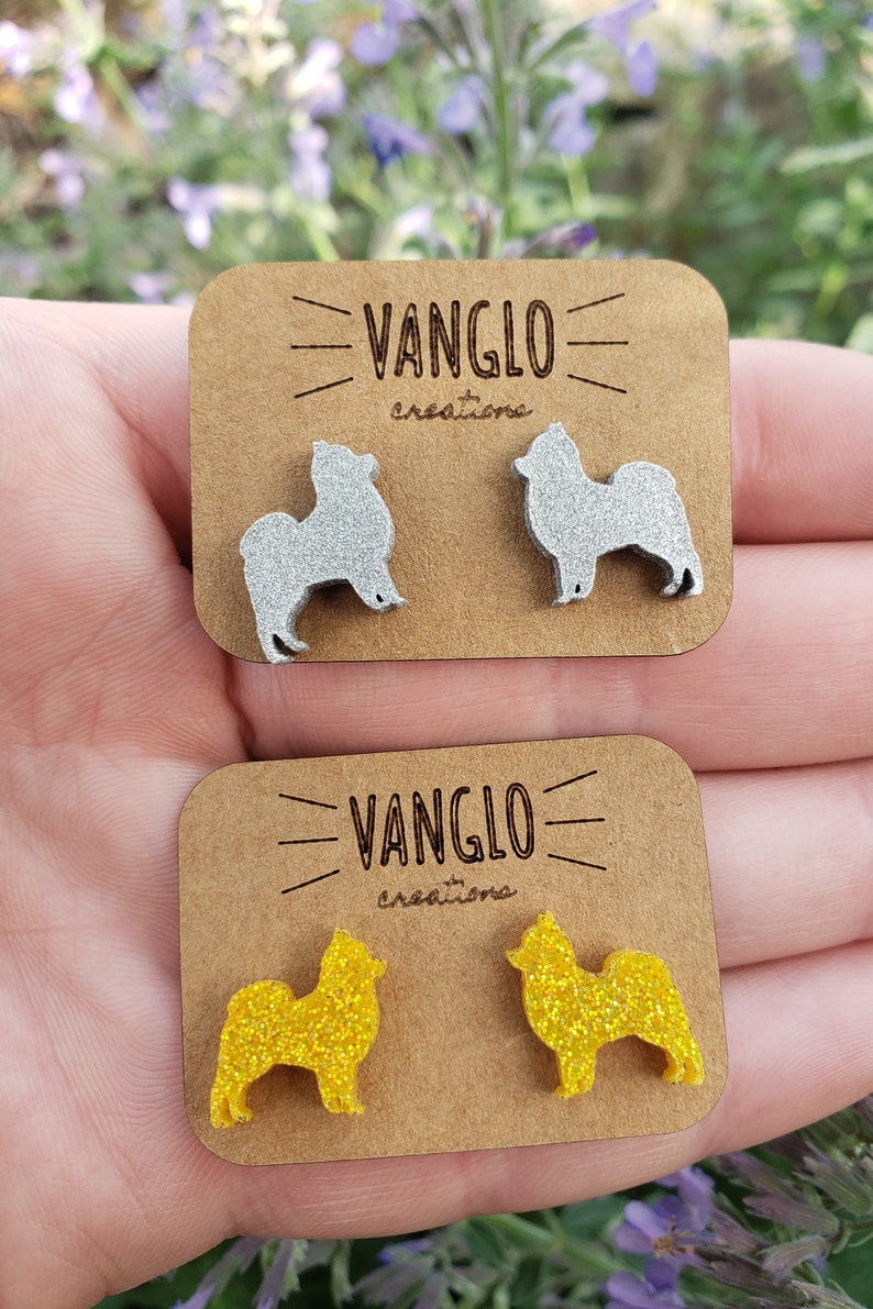 May include: Two pairs of dog-shaped earrings. One pair is silver glitter and the other is yellow glitter. The earrings are on a brown card with the text "VANGLO creations" printed on it.