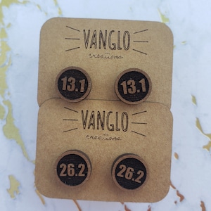Puede incluir: Cuatro pendientes redondos de madera con letras negras. Dos pendientes dicen "13.1" y dos pendientes dicen "26.2". Los pendientes se muestran en una tarjeta marrón con el texto "VANGLO Creations" impreso en ella.