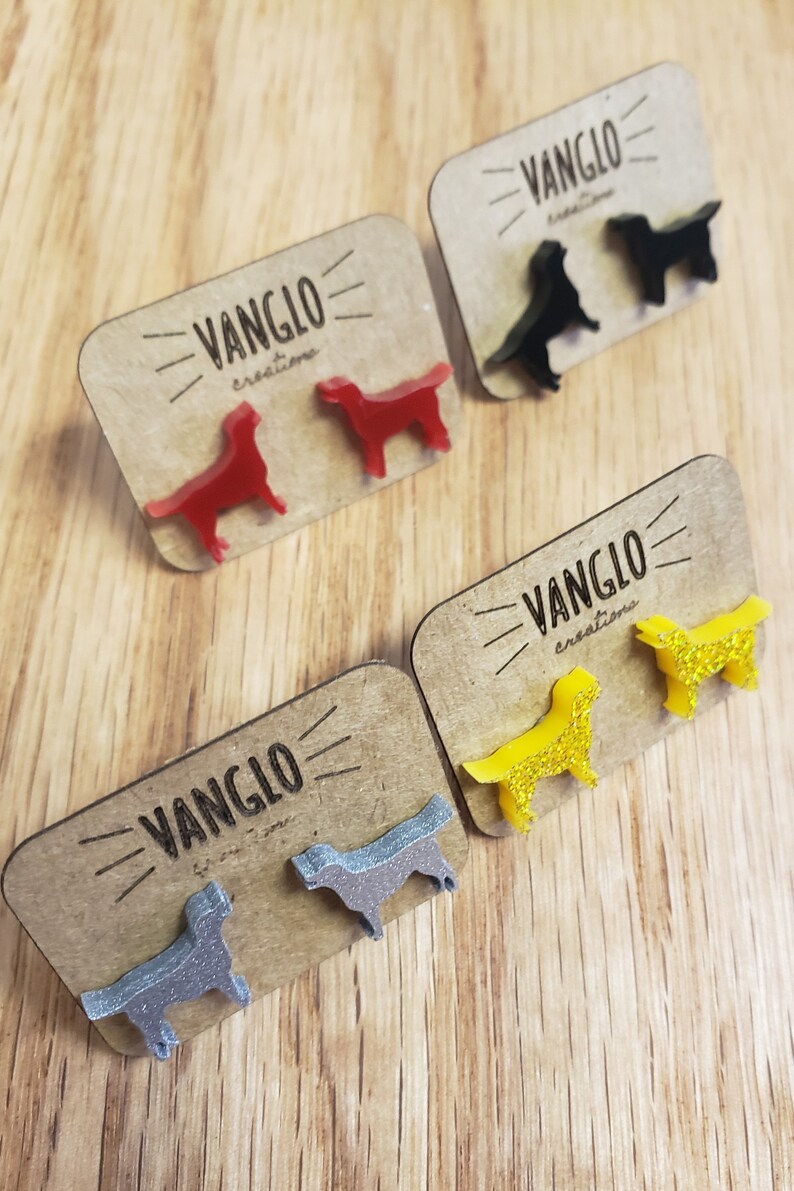 May include: Four pairs of dog-shaped earrings on display cards. The earrings are made of acrylic and come in red, black, yellow, and silver. Each card has the text "VANGLO creations" printed on it.
