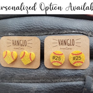 Puede incluir: Dos pares de pendientes con temática de softball. Un par presenta un softball en forma de corazón con costuras rojas y el otro par presenta un softball redondo con el número 25 impreso en él. Ambos pares están en una tarjeta marrón con el texto "VANGLO Creations" impreso en ella.