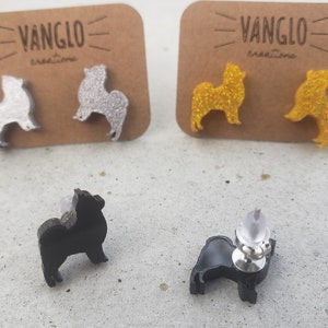 May include: A pair of silver glitter, gold glitter, and black dog silhouette earrings. The earrings are on a white surface with a brown card behind them. The card has the text "VANGLO creations" on it.