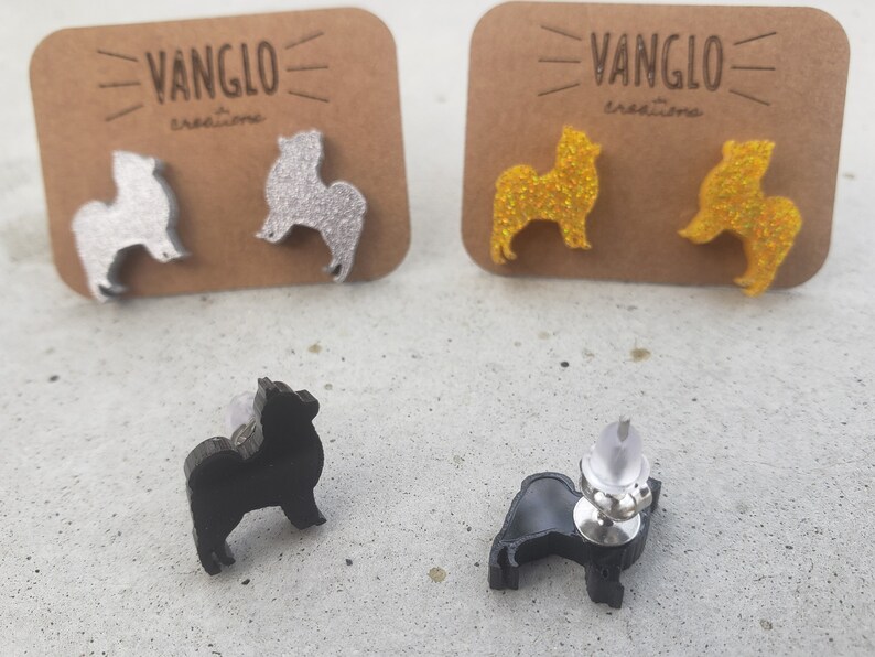 May include: A pair of silver glitter, gold glitter, and black dog silhouette earrings. The earrings are on a white surface with a brown card behind them. The card has the text "VANGLO creations" on it.