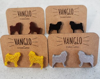 May include: Four pairs of pug dog earrings on display cards. The earrings are made of glitter and come in four colors: brown, black, gold, and silver. The cards are brown and have the text "VANGLO creations" printed on them.